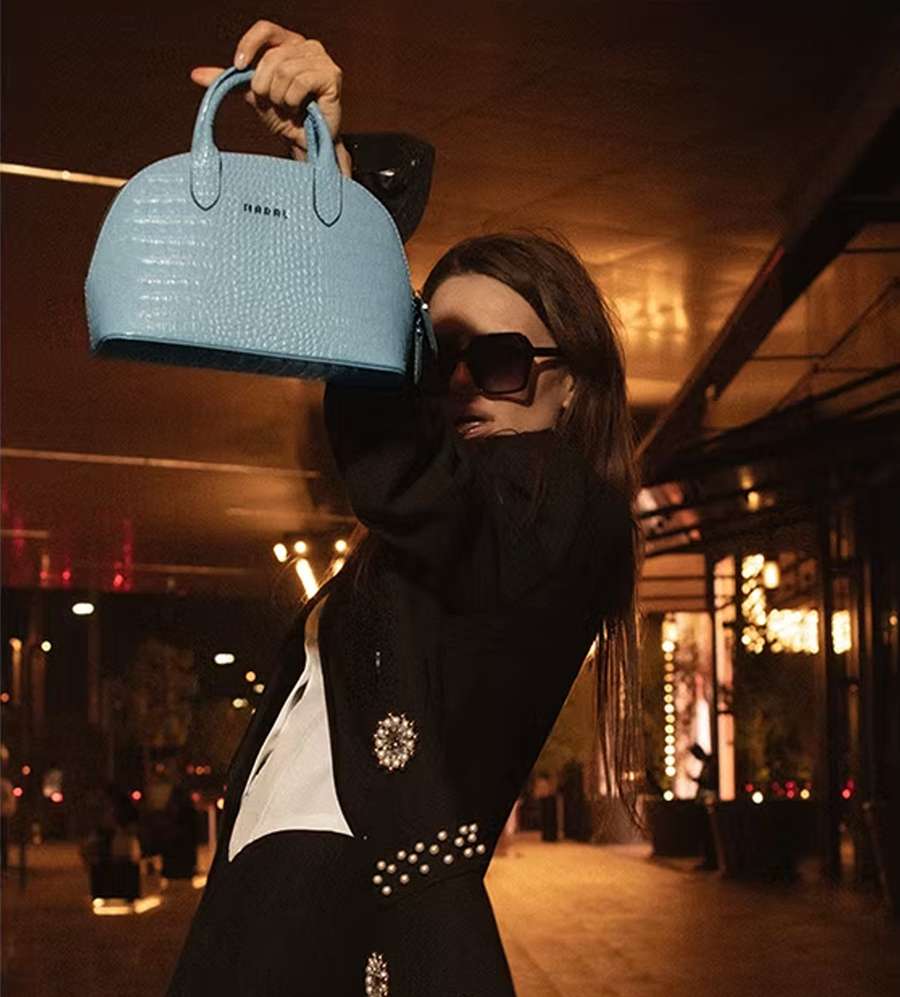 A woman in sunglasses and a black jacket with jewelled buttons, holding up a light blue handbag on a street at night