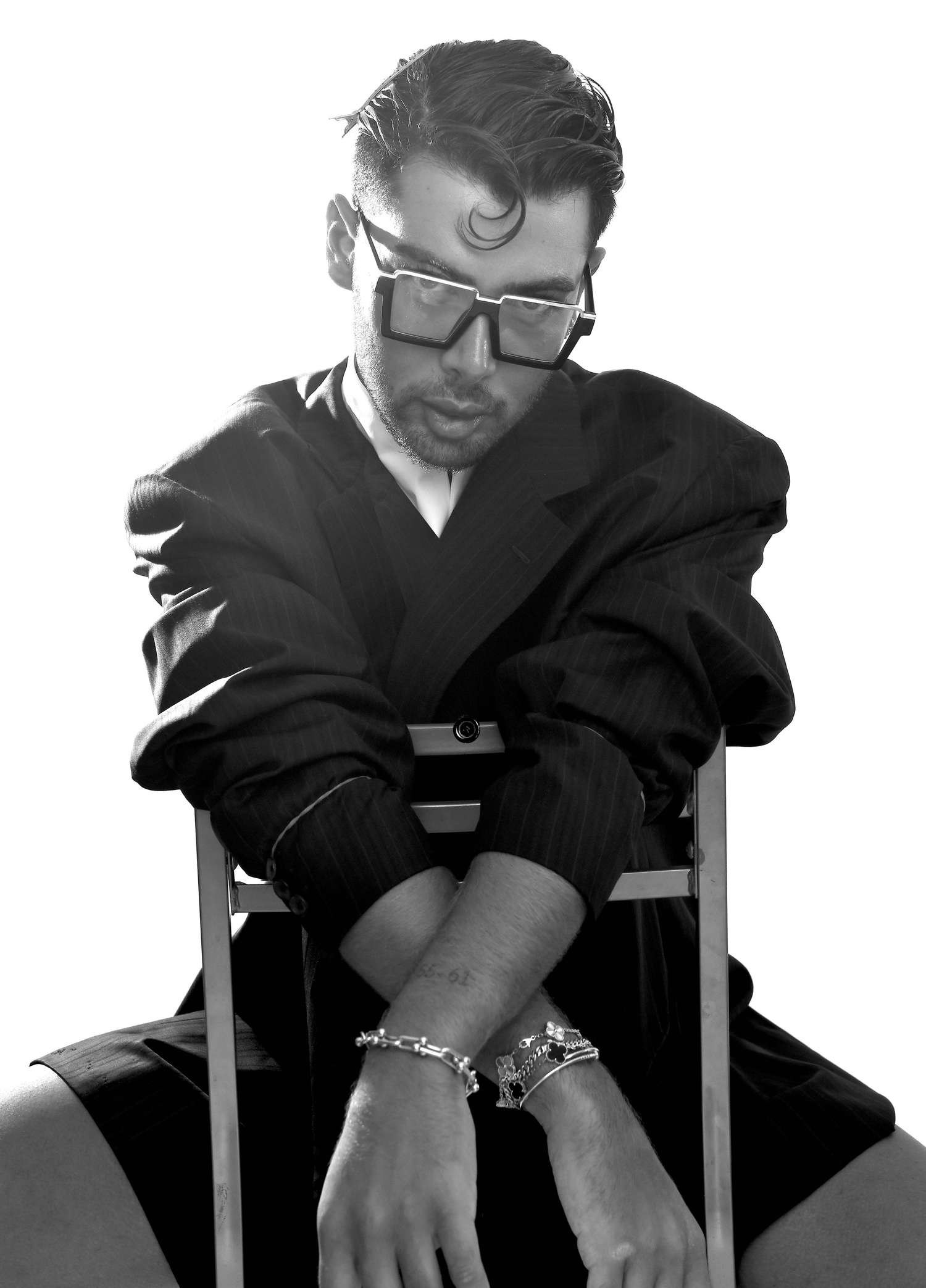 Black and white portrait of a man in square glasses and a pinstripe jacket, leaning on the back of a chair with his wrists crossed