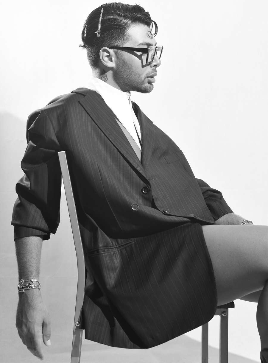 Black and white side view of a man in an oversized pinstripe jacket and square glasses, sitting on a chair