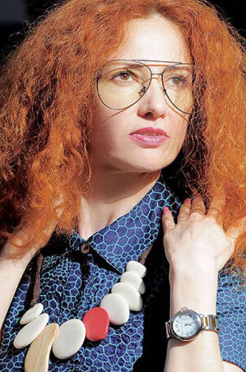 Portrait of a woman with curly red hair and clear aviator glasses, wearing a blue patterned shirt and a large necklace of white and red discs